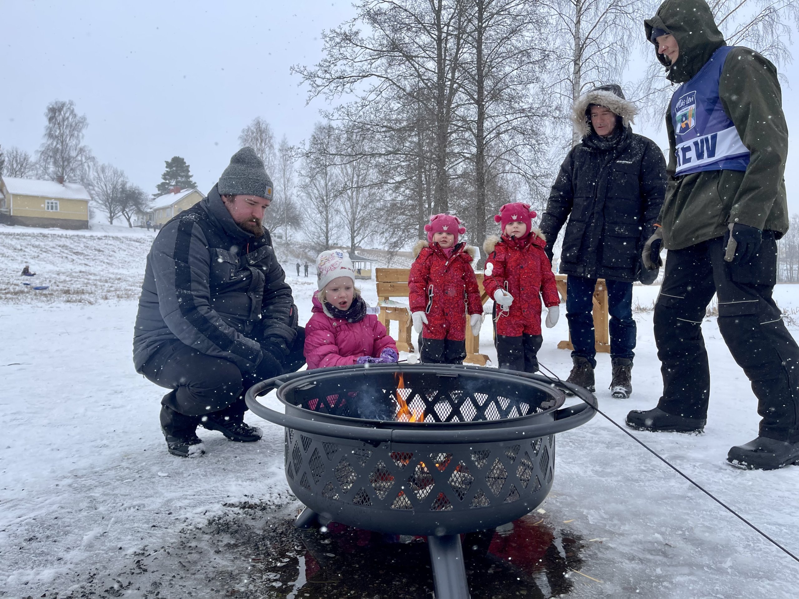 Kolme pientä tyttöä ja kaksi miestä paahtavat vaahtokarkkeja talvimaisemassa nuotion ääressä.